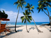 Beach scene with clear blue sky and ocean, palm trees, white sand, and a red lifeguard tower. Few people are relaxing near the shoreline.