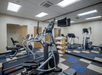 A gym room with treadmills, exercise bikes, and a rowing machine on a checkered carpet floor. A wall-mounted TV and mirror are visible.