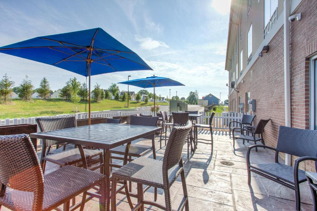 Outdoor patio with wicker chairs and tables under blue umbrellas on a sunny day, adjacent to a brick building and grassy area.