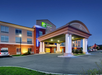 Exterior of a three-story Holiday Inn Express hotel at dusk, featuring a covered entrance, illuminated signage, parked cars, and surrounding greenery.