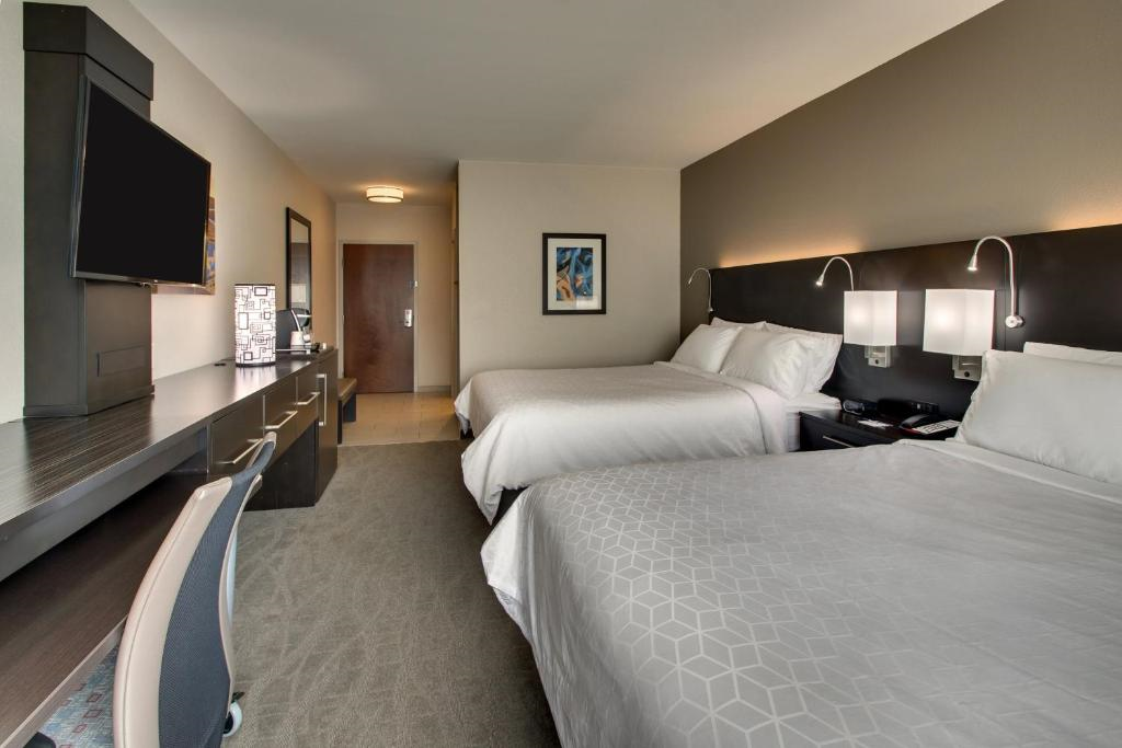 A modern hotel room with two double beds, a wall-mounted TV, desk, chair, and lamps. The room has neutral colors and a window in the background.
