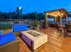 Outdoor seating area with wicker furniture, blue cushions, a gas fire pit table, and a wooden pergola with string lights on a deck. Trees and a fence are in the background.