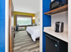 A hotel room with a neatly made bed, a window, a desk, a chair, a small fridge, a microwave, and a coffee maker on a shelf. Wall colors are blue and yellow.