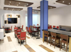 A modern dining area with red and brown chairs, tables, blue pillars, and a wall-mounted TV. The room features overhead lighting and a tiled floor.