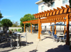 Outdoor patio with wooden pergola, tables, and chairs under umbrellas. Bright and sunny day with trees and a building in the background.
