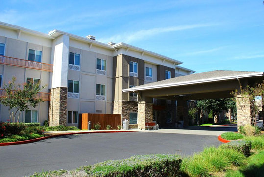 Three-story hotel building with a neutral color scheme, featuring balconies and stone accents, surrounded by landscaping and a driveway.