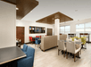 A dining area with red, blue, cream, and green chairs at gray tables and floor to ceiling windows letting light in.