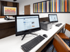 A small business center with two computers on a long white desk with a printed between them and two brown chairs in front of them.