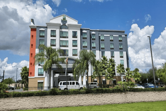 The white and gray exterior of the Holiday Inn Express & Suites with a line of palm tress in front of it on a sunny day in Orlando, Florida.