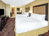 Hotel room with two large beds, white bedding, a TV, a dresser, and a kitchenette. Walls are beige and carpet is patterned. The room looks tidy and well-lit.