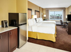 Hotel room with two double beds, a kitchenette with a mini-fridge and a coffee maker, a desk, TV, and a window with a view of neighboring buildings.