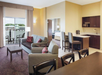 A hotel suite with a living area, sofa, armchair, and coffee table. Nearby are a kitchenette with a counter and a separate bedroom with a bed visible through a doorway.