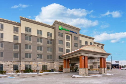 Four-story Holiday Inn Express & Suites hotel with stone and beige exterior, covered entrance, and parking lot under a partly cloudy sky.