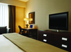 Hotel room with a bed, a TV on a dresser, and a desk with a chair and lamp. A window with curtains is on the left.