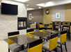 Modern dining area with yellow and black chairs, granite tables, a wall-mounted fireplace, and a TV. Neutral-toned walls are decorated with framed art and a clock.