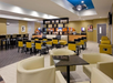 Modern hotel dining area with tables, chairs, and a breakfast buffet station. Bright lighting and contemporary decor.