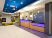 Hotel lobby with modern decor, blue accents, and a reception desk with a staff member behind it. Yellow seating and a fireplace visible in the background.