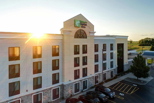 Exterior of a four-story hotel building with a Holiday Inn Express & Suites sign, seen during a sunset. The parking lot is visible in the foreground.