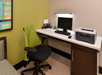 Office desk with a computer, keyboard, and a Brother printer. A green mesh office chair is positioned nearby. The wall has light green and beige colors with a framed abstract artwork.
