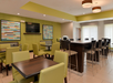 A modern, empty hotel dining area with green chairs, wood tables, a wall-mounted TV, abstract art, and ceiling lights.