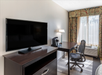 Hotel room with a flat-screen TV on a dresser, a desk with a chair, a lamp, and a coffee maker. Window with patterned curtains in the background.