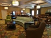 A hotel breakfast area with tables, chairs, a sofa, and a coffee table. Beverage machines and breakfast supplies are in the background, with decorative ceiling lights above.