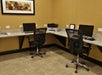 Two office chairs at a corner desk with three computers and a printer in a room with beige walls and a patterned floor. A framed abstract painting hangs above the desk.
