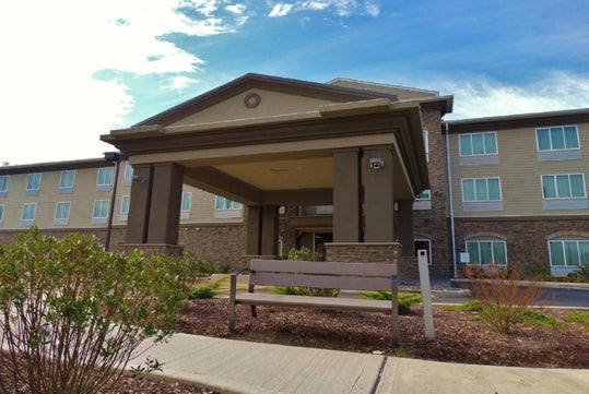 Modern hotel entrance with a covered driveway, brown brick and beige facade, and a bench in the foreground, surrounded by small shrubs under a partly cloudy sky.