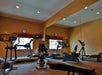 A gym room with treadmills, exercise bikes, mirrors, and wall-mounted TVs. The carpet is gray, and the walls are beige. Clocks are visible on the wall.