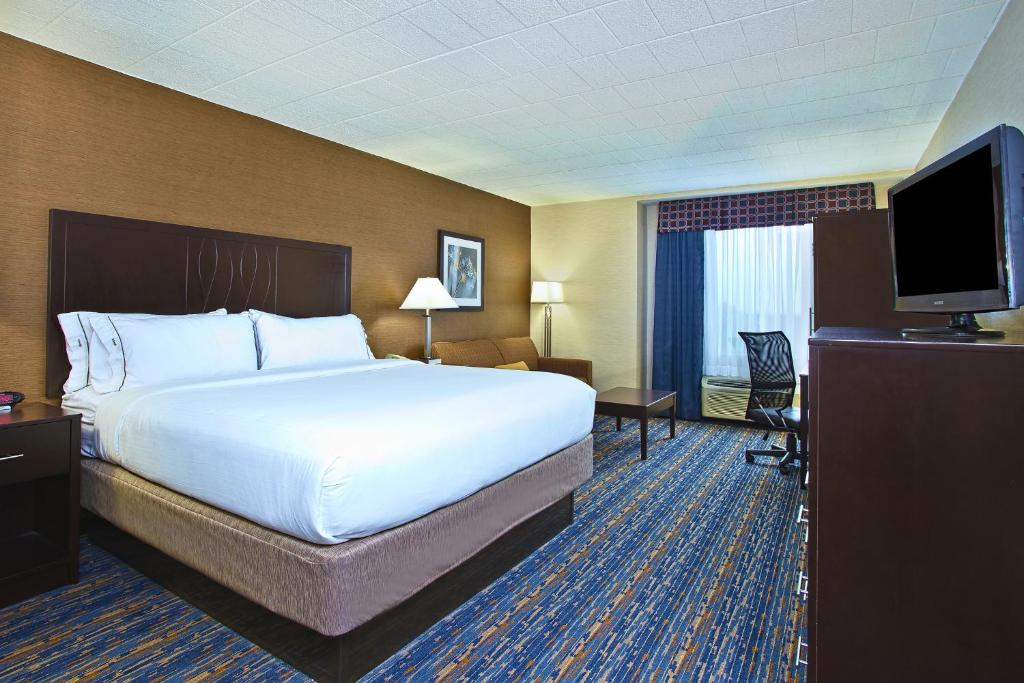 Hotel room with a large bed, bedside table, sofa, floor lamp, TV on a dresser, desk with chair, and window with curtains. Carpeted floor and neutral wall colors.