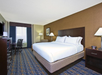 A hotel room with a large bed, two bedside lamps, a TV, work desk with chair, and a window with curtains. Brown and beige decor with blue accents.