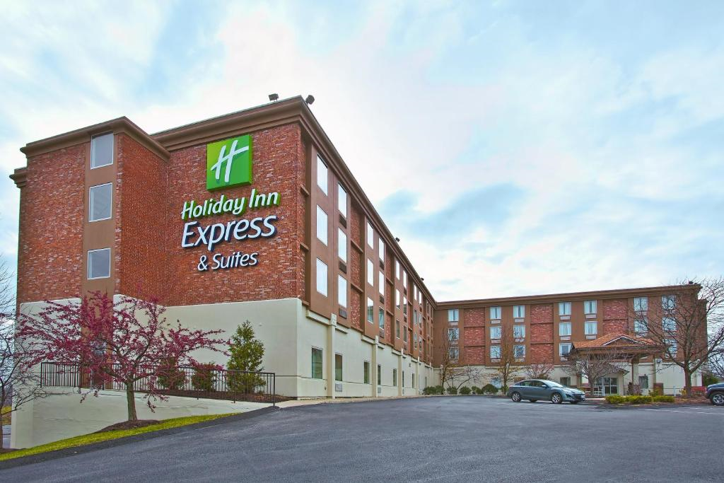 A brick and cream-colored Holiday Inn Express & Suites building with green signage, set against a cloudy sky. A parking lot with a few cars is visible in the foreground.