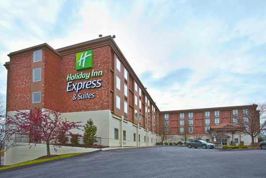 A brick and cream-colored Holiday Inn Express & Suites building with green signage, set against a cloudy sky. A parking lot with a few cars is visible in the foreground.