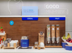 Breakfast buffet with sections for cereal and fruit. Oatmeal pot, cereal dispensers, fruit bowl, and condiments like sriracha are displayed. Sign reads "GOOD MORNING.