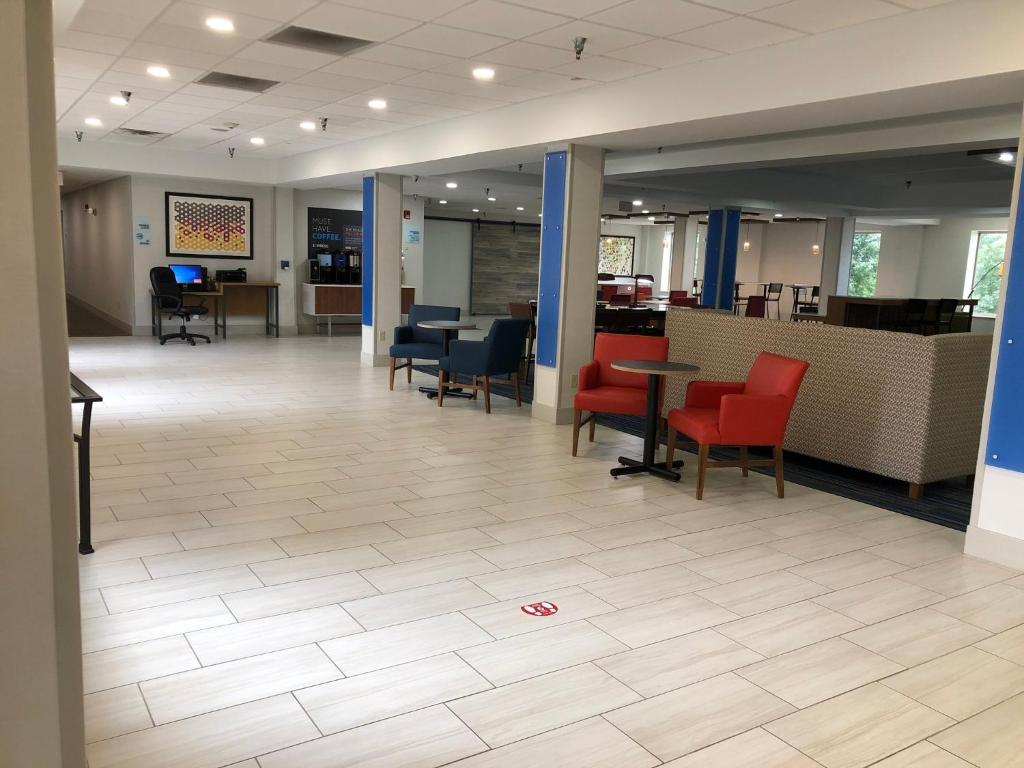Lobby area with tiled floor, red and blue seating, tables, and a computer station. Walls are decorated with framed art. Bright lighting and large windows provide natural light.