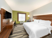 Hotel room with a large bed, wooden headboard, desk, chair, wall-mounted TV, and a window with soft light. Walls are white and green. Floor has a striped carpet.