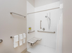 Accessible shower with grab bars, a handheld showerhead, a foldable bench, and three folded towels on a wall rack.