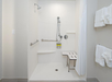 A white accessible shower with a detachable showerhead, grab bars, a shower bench, and toiletries on a small shelf. White towels hang on the wall to the right.