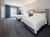 Hotel room with two double beds, modern decor, and a window with a blind. Carpeted floor, nightstand with a phone, and a wall lamp.