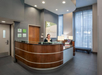A receptionist sits behind a curved wooden desk in a modern hotel lobby with gray walls, large windows, and a Holiday Inn sign.