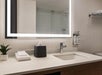 Modern bathroom with a lighted mirror, sink, tissue box, folded towels, and mounted soap dispensers on the right wall. A small plant is on the counter.