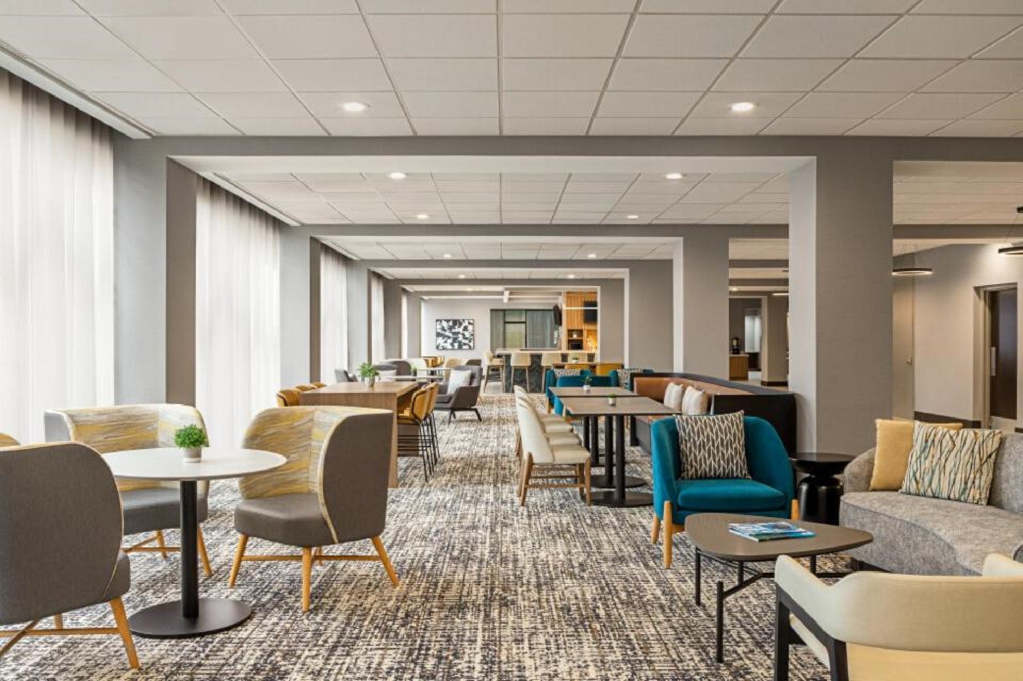 Modern lounge area with various seating arrangements, tables, and soft lighting, featuring neutral tones and patterned carpet floors.