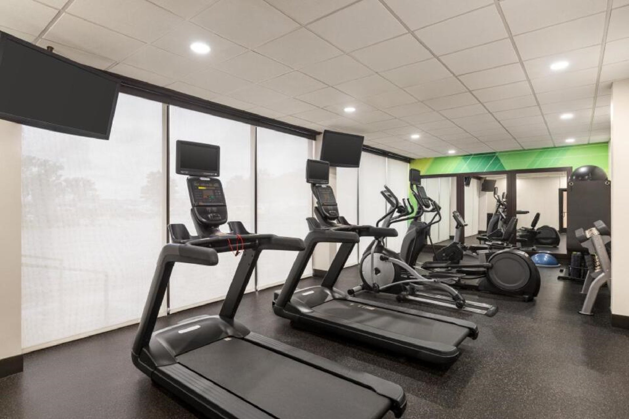 A gym with treadmills, elliptical machines, and various exercise equipment. The room has several mounted screens and large windows with blinds.