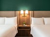 Hotel room with two neatly made double beds, a shared nightstand between them, a small plant, a book, and a wall-mounted lamp above.