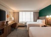 Hotel room with two double beds, a desk with a chair, a lamp, a TV on a cabinet, and a window with curtains. Walls are green and white with framed art. Room is neatly arranged.