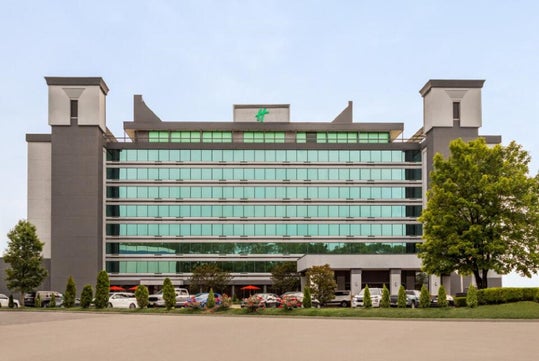 A multi-story hotel with a modern facade, featuring numerous windows and two prominent towers on either side, is bordered by trees and a parking area with parked cars.