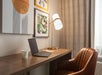 A modern desk with a laptop, cup, and lamp, flanked by a brown chair. Abstract artwork hangs on the wall, and beige curtains are partially opened.