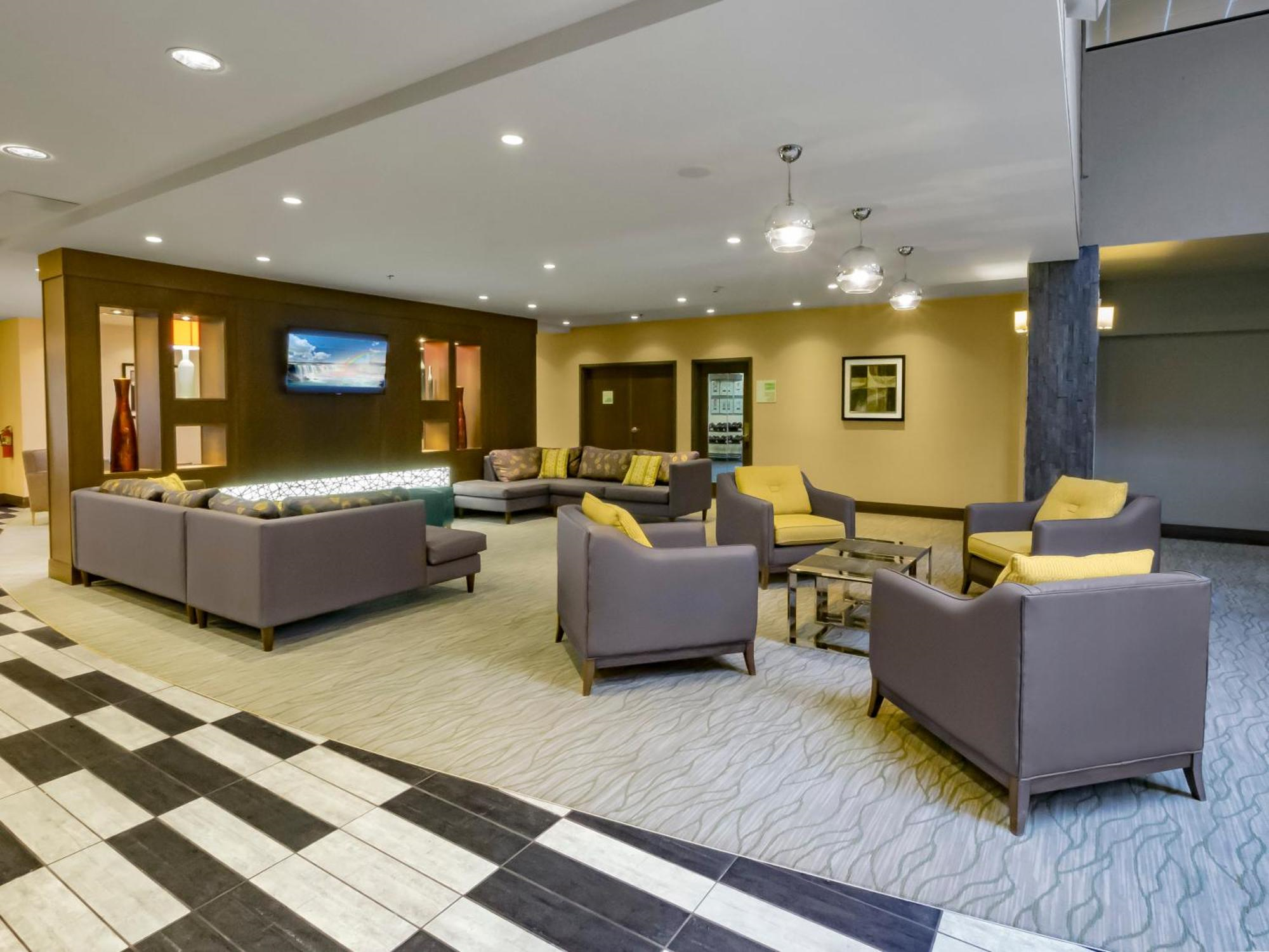 Modern hotel lobby with brown and yellow chairs, several couches, a wall-mounted TV, and overhead lighting. The floor has patterned carpet and tile.