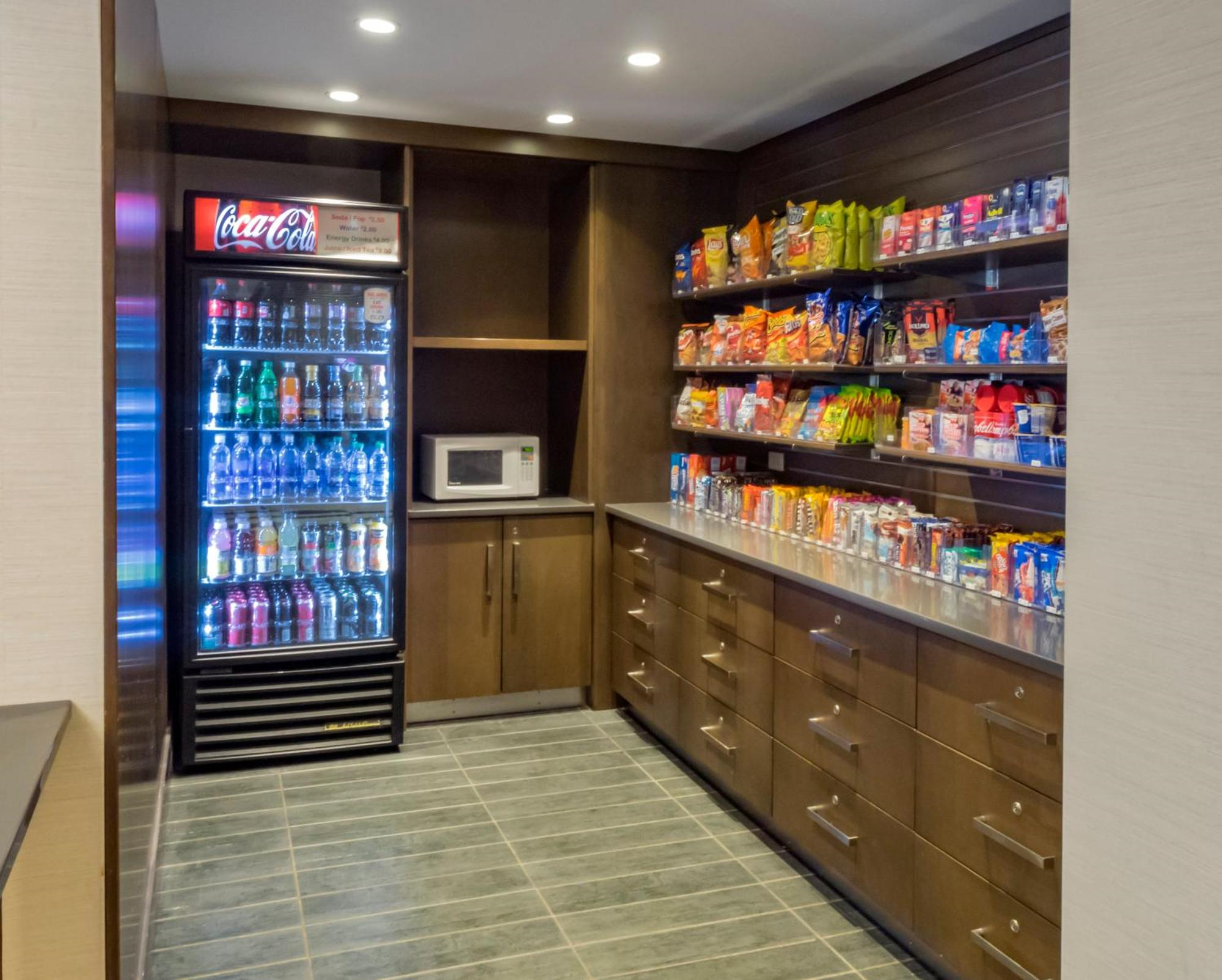 Small convenience store area with a refrigerated drinks display, microwave, and shelves stocked with snacks and beverages, set against wood cabinets and tile flooring.