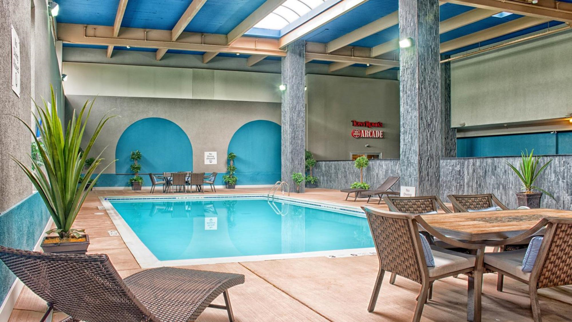 Indoor swimming pool with blue water, surrounded by tables, chairs, potted plants, and columns under a skylight ceiling.